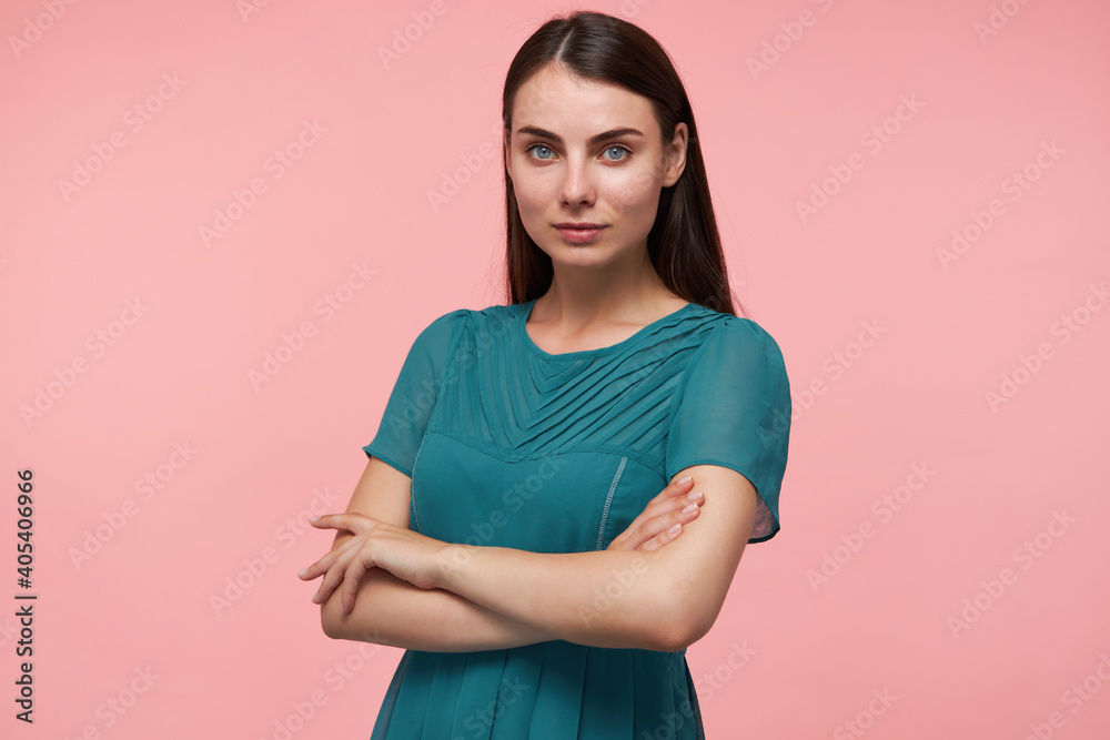 Young lady, pretty woman with long brunette hair. Folding hands on a chest, crossing hands and watching at the camera isolated over pastel pink background. Wearing emerald dress Young lady, pretty woman with long brunette hair. Folding hands on a chest, crossing hands and watching at the camera isolated over pastel pink background. Wearing emerald dress