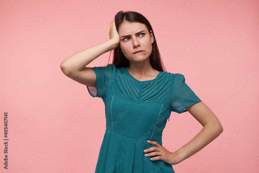 Young lady, pretty woman with long brunette hair. Holding one hand on a waist and touching her head, thinking. Watching to the right at copy space over pastel pink background. Wearing emerald dress Young lady, pretty woman with long brunette hair. Holding one hand on a waist and touching her head, thinking. Watching to the right at copy space over pastel pink background. Wearing emerald dress