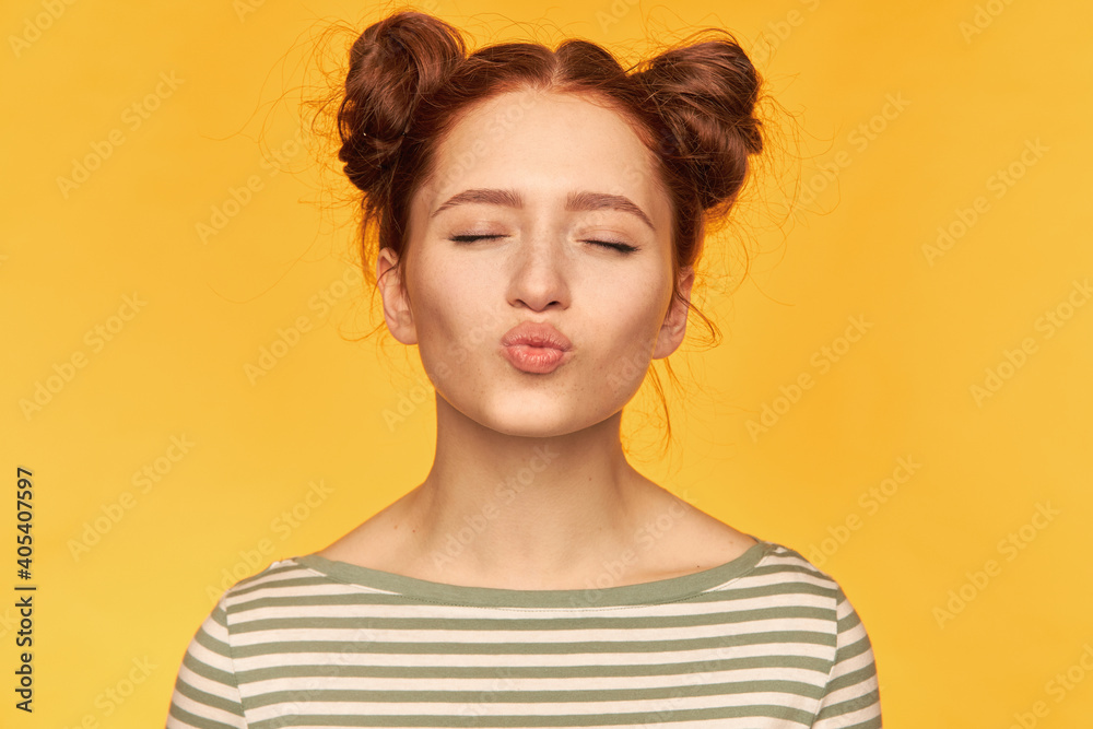 Portrait of attractive, lovely, red hair girl with two buns and healthy skin. Sweet kiss with closed eyes. Wearing striped sweater and stand isolated, closeup over yellow background Portrait of attractive, lovely, red hair girl with two buns and healthy skin. Sweet kiss with closed eyes. Wearing striped sweater and stand isolated, closeup over yellow background