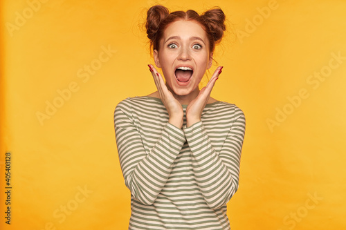 Young lady, horrified looking ginger woman with two buns. Shows how much she frightened of what she see. Wearing striped sweater and screams at the camera isolated over yellow background