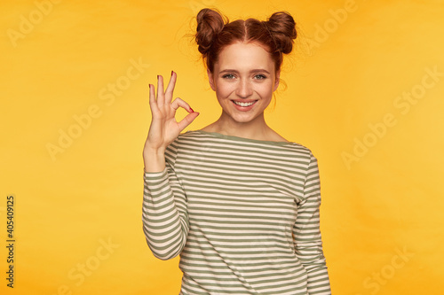 Teenage girl, happy, successful looking red hair woman with two buns. Wearing striped sweater and showing okay sign, smile. Watching at the camera isolated over yellow background