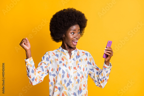Photo of shiny lucky dark skin lady dressed casual outfit looking holding modern device rising fist isolated yellow color background