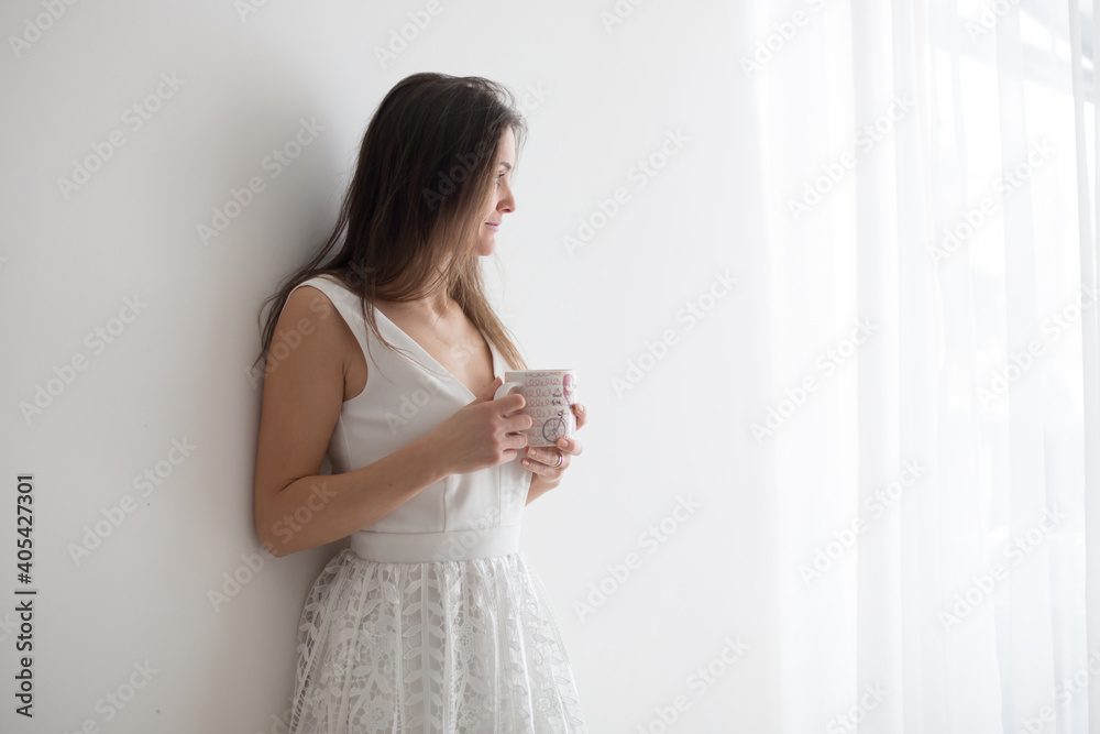 Middle age woman with white dress, drinking coffee at home, looking out of the window Middle age woman with white dress, drinking coffee at home, looking out of the window