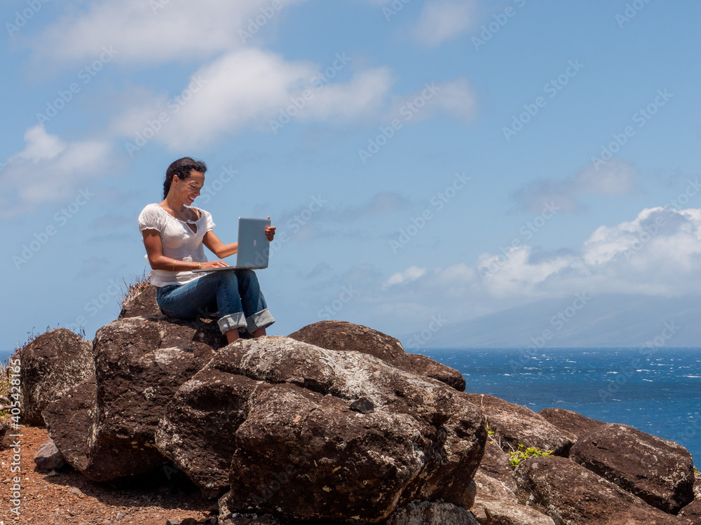 A woman sitting on a rock overlooking the ocean working on her laptop