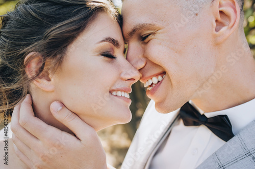 Close-up faces of a groom in a gray suit, a black bow tie, a boutonniere and a beautiful brunette bride in a white dress with curly hair on a nature background.