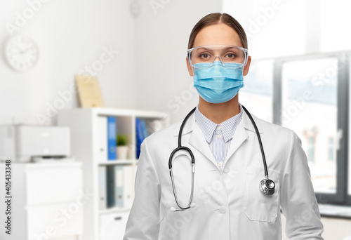 health, medicine and pandemic concept - young female doctor with stethoscope wearing goggles and face protective medical mask for protection from virus disease over hospital background
