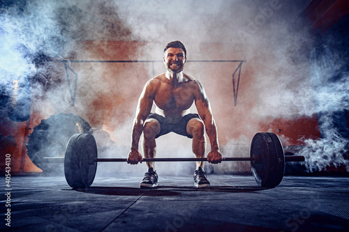 Muscular fitness man doing deadlift a barbell over his head in modern fitness center. Functional training. Snatch exercise. Smoke on background.