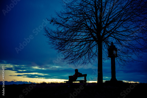 silhouette of a mysterious hacker. Silhouette portrait of a lonely anonymous man sitting on a bench as dark clouds above him roll, with the sun beginning to set behind 