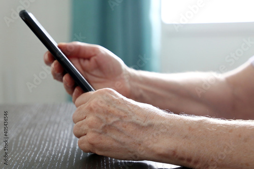 Elderly woman with smartphone sitting at the table  mobile phone in wrinkled female hands. Concept of online communication at retirement