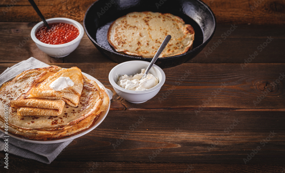 Traditional Russian Crepes Blini stacked in plate and cast-iron frying pan with red caviar, fresh sour cream on dark wooden table. Russian festival meal Maslenitsa or Shrovetide. Space for text Traditional Russian Crepes Blini stacked in plate and cast-iron frying pan with red caviar, fresh sour cream on dark wooden table. Russian festival meal Maslenitsa or Shrovetide. Space for text