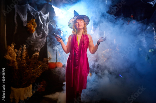 Blonde witch in red dress and black hat in Halloween decoration indoors with cell phone and smoke near her. Woman taking a selfie during holiday carnival