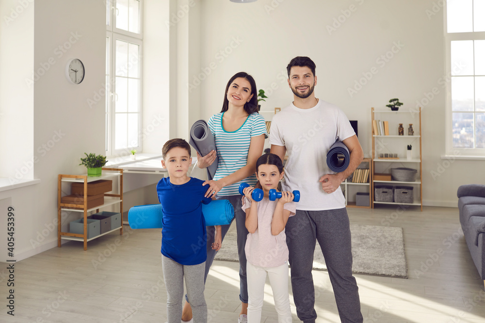 Fototapeta premium Active family sports workout in lockdown at home concept: Portrait of happy fit young couple with healthy little children standing together, holding exercise mats and dumbbells and looking at camera