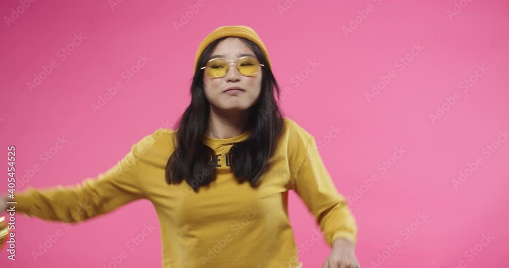Portrait of young Asian positive pretty young woman in yellow stylish glasses dancing to music or song having fun moving rhythmically holding golden heart shape balloon isolated on pink wall in studio Portrait of young Asian positive pretty young woman in yellow stylish glasses dancing to music or song having fun moving rhythmically holding golden heart shape balloon isolated on pink wall in studio