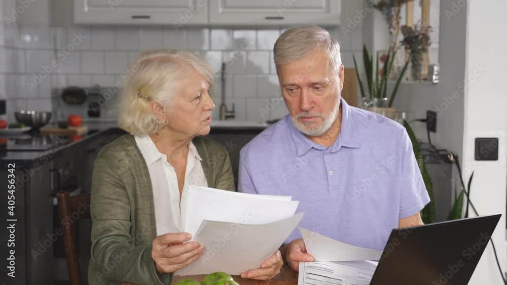 A sixty-year-old wife and husband sit at a desk and laptop, holding bills in their hands, managing the family budget, checking utility bills. A sixty-year-old wife and husband sit at a desk and laptop, holding bills in their hands, managing the family budget, checking utility bills.