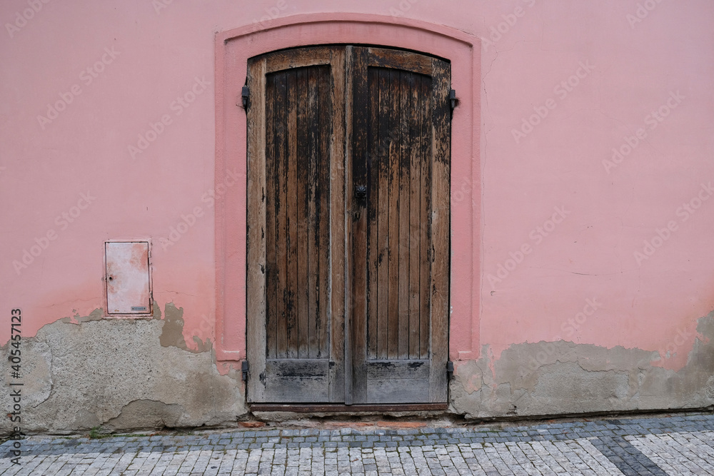 Fototapeta premium Vintage wooden painted door of old building. Shabby door boards. Old wall made of pink plaster and gray putty stains on the wall. Smooth surface. Cracks. Old peeling plaster wall, crumbles.