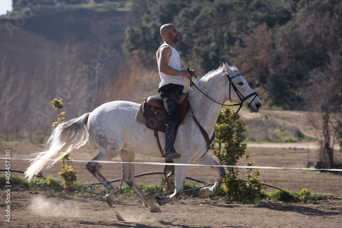 Young guy in casual outfit riding white horse on sandy ground