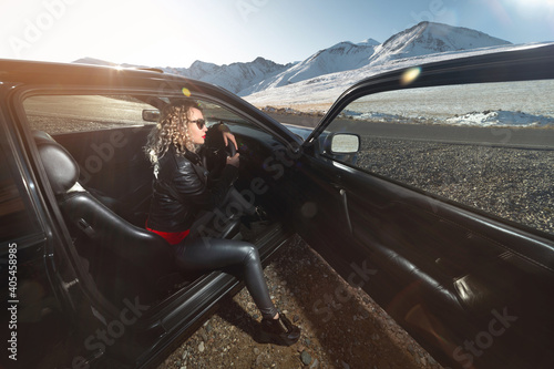 An attractive blonde in sunglasses and leather clothes sits in a black leather salon with an open door of a sports car on a country road in the mountains at sunset. Retro style 90s © yanik88