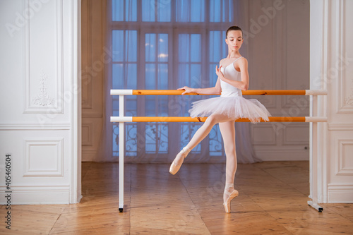 young slender ballerina in a white tutu standing on pointe at ballet bar in a beautiful white hall in front of a mirror.