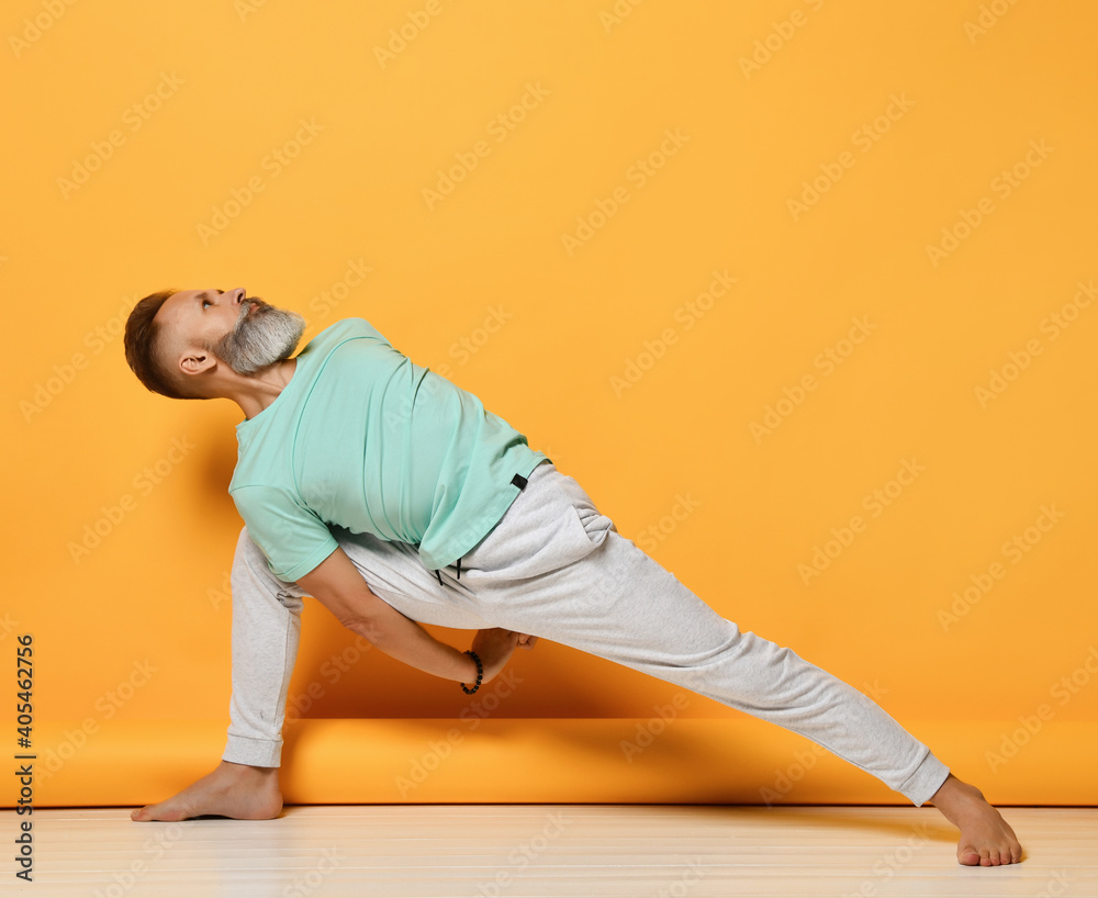 Active barefoot mature handsome grey-haired man practicing yoga stretching back and arm in warrior asana holding balance. Stability and strength achievement. Fitness pilates and workout