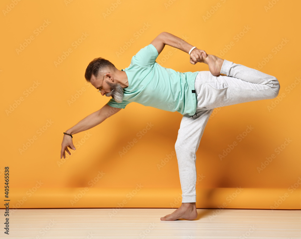 Healthy gray brutal bearded man wearing glasses casual clothes balancing on one leg, stretching the muscles, practicing fitness or yoga exercises for Pilates. Isolated portrait on orange background Healthy gray brutal bearded man wearing glasses casual clothes balancing on one leg, stretching the muscles, practicing fitness or yoga exercises for Pilates. Isolated portrait on orange background