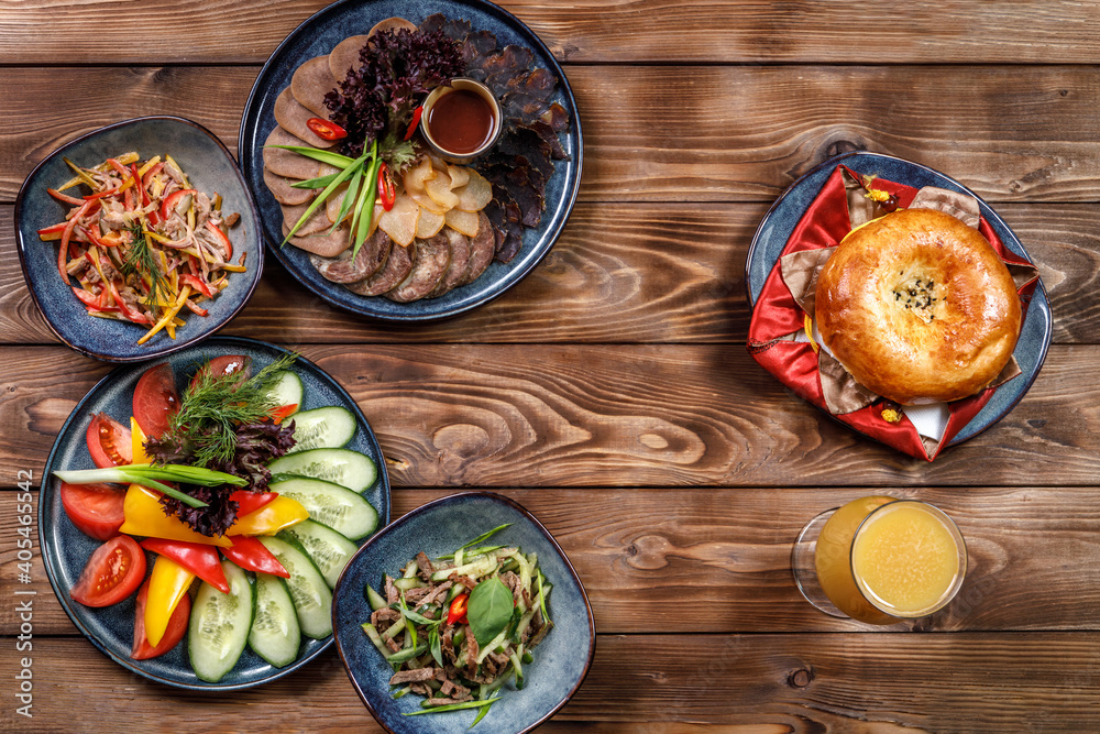 Flat layout of various dishes with sliced vegetables, meat cuts, salads, orange juice and bun on a brown wooden background.