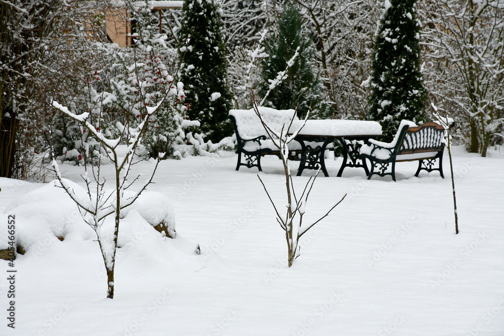 Close up on a set of chairs and tables located in the garden that are covered with snow seen next to a chimney, a set of small trees and decorative shrubs and other vegetation spotted in winter