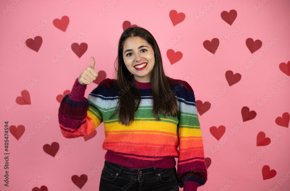 Young beautiful woman over pink background with harts doing positive gesture with hand, thumbs up smiling and happy. cheerful expression and winner gesture. Young beautiful woman over pink background with harts doing positive gesture with hand, thumbs up smiling and happy. cheerful expression and winner gesture.