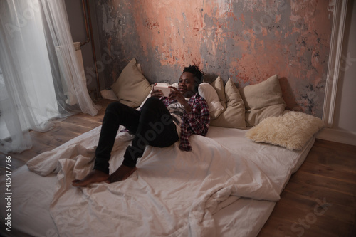 young african american man lies on the bed with headphones looking at the phone