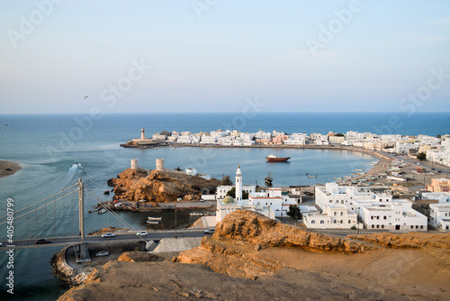 Aerial view of the village of Al Ayjah from the top of a mountain. There are boats on the sea, a suspension bridge and white buildings along the bay. Sur, Oman.