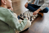 Little thoughtful boy playing chess with grandfather while sitting on the floor in living room at home