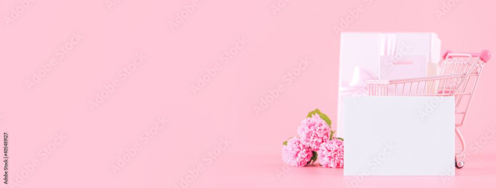 Shopping cart and gift with carnation on pink table background.