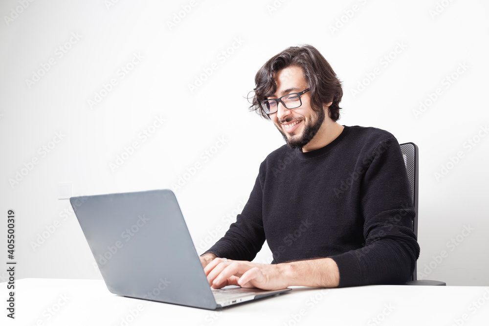 Middle eastern adult bearded man with long hair and glasses using laptop computer while smiles on a white office room with copy space. Remote working concept.