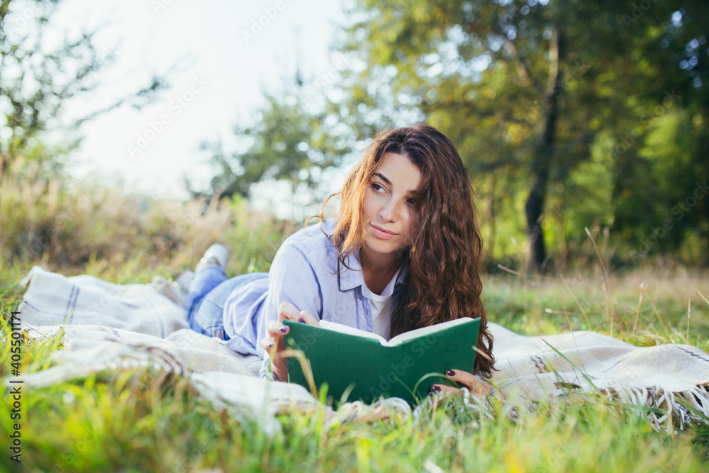 Beautiful young woman with curly black hair, resting lying on a mat in the park on the grass, and holding a book reads Beautiful young woman with curly black hair, resting lying on a mat in the park on the grass, and holding a book reads