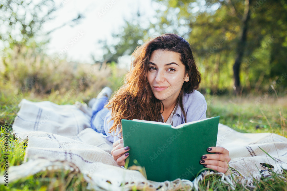 Obraz premium Beautiful young woman with curly black hair, resting lying on a mat in the park on the grass, and holding a book reads