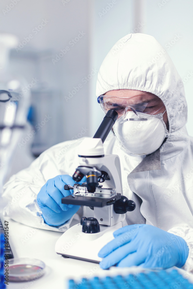 Medical engineer adjusting microscope while doing coronavirus investigation Scientist in protective suit sitting at workplace using modern medical technology during global epidemic. Medical engineer adjusting microscope while doing coronavirus investigation Scientist in protective suit sitting at workplace using modern medical technology during global epidemic.