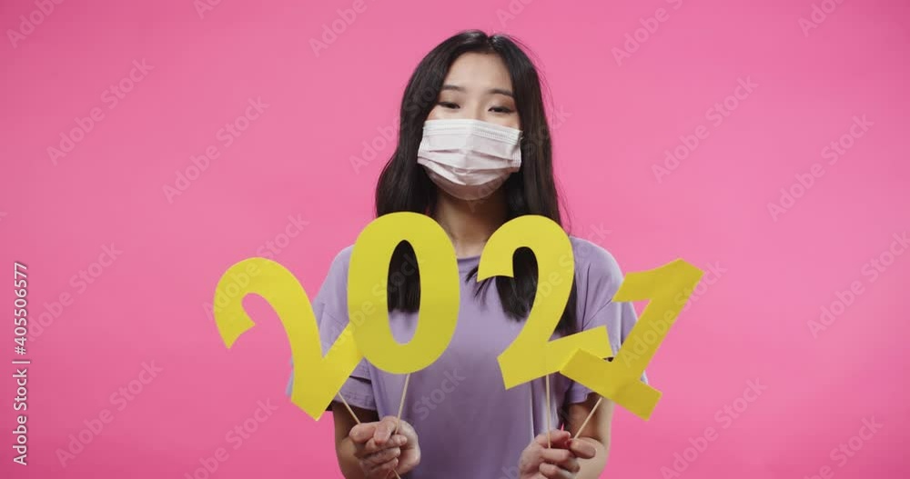Portrait of joyful Asian young woman teen in mask smiling and looking at camera standing isolated on pink background holding 2021 sign in hands, New Year celebration in quarantine Portrait of joyful Asian young woman teen in mask smiling and looking at camera standing isolated on pink background holding 2021 sign in hands, New Year celebration in quarantine