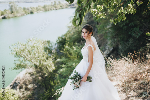 Wallpaper Mural Wedding portrait of a cute bride with a bouquet in a long white dress and curly hair on a cliff, against the backdrop of nature with green trees. Torontodigital.ca
