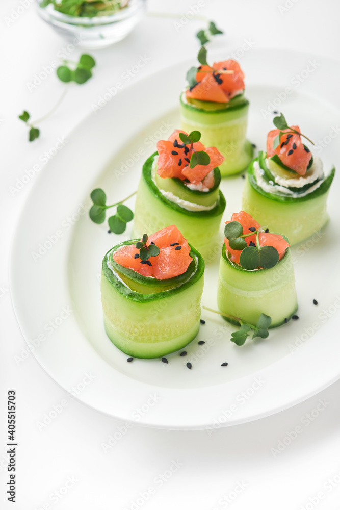 Holiday vegetable appetizers. Cucumbers rolls with soft cheese, pieces of salted salmon, microgreens and black sesame served on a white plate. Selective focus. Holiday vegetable appetizers. Cucumbers rolls with soft cheese, pieces of salted salmon, microgreens and black sesame served on a white plate. Selective focus.