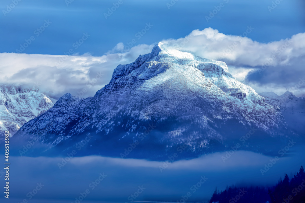 Majestic mountain peak covered in snow with a blanket of fog around its base and beautiful clouds in background, Glacier National Park, Montana