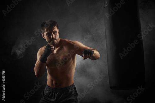 sporty shirtless boxer in black boxing wraps punching in boxing bag on dark background with smoke