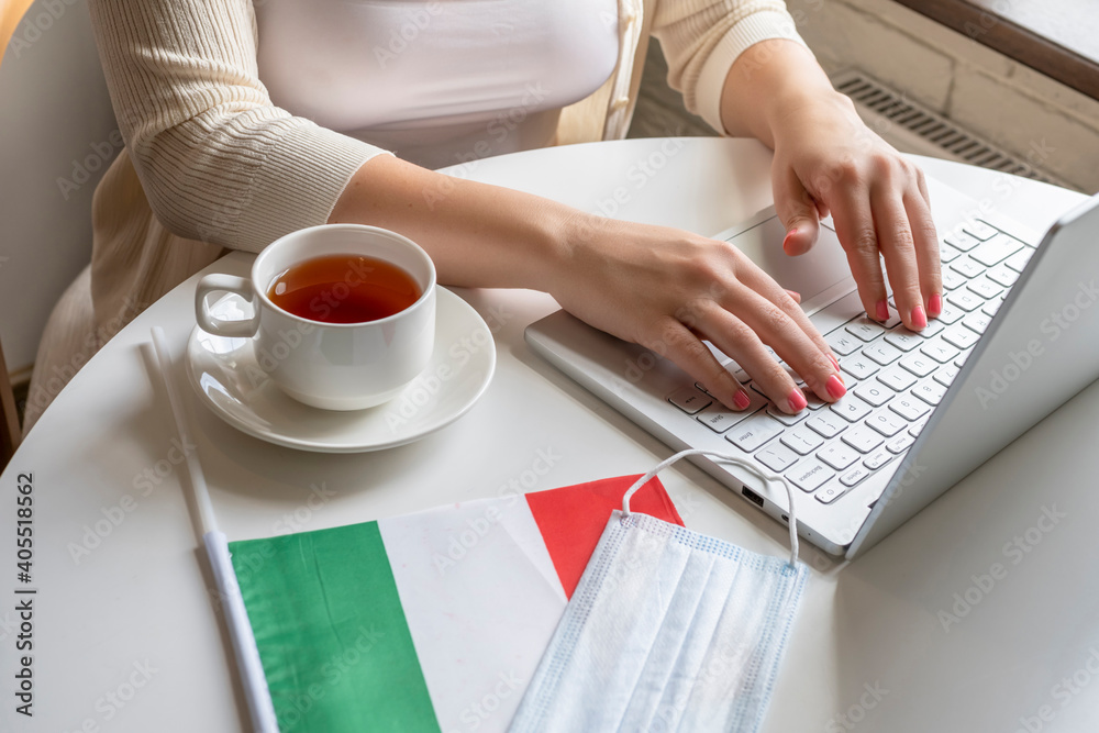 Woman tourist having breakfast with cup of coffee working on laptop. Flag of Italy, medical protective face mask on table of cafe. Protection from bacteria and viruse in a public place. Concept. Woman tourist having breakfast with cup of coffee working on laptop. Flag of Italy, medical protective face mask on table of cafe. Protection from bacteria and viruse in a public place. Concept.
