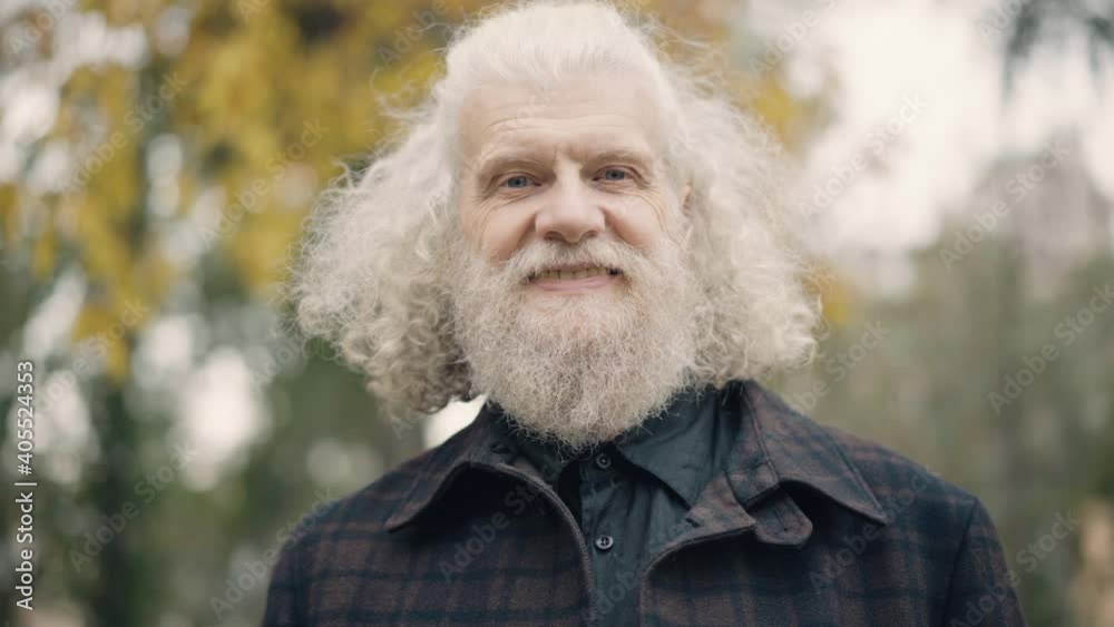 Close-up of senior man looking away smiling and turning to camera. Headshot of confident handsome Caucasian male retiree with long grey hair posing in autumn park. Happiness concept. Close-up of senior man looking away smiling and turning to camera. Headshot of confident handsome Caucasian male retiree with long grey hair posing in autumn park. Happiness concept.