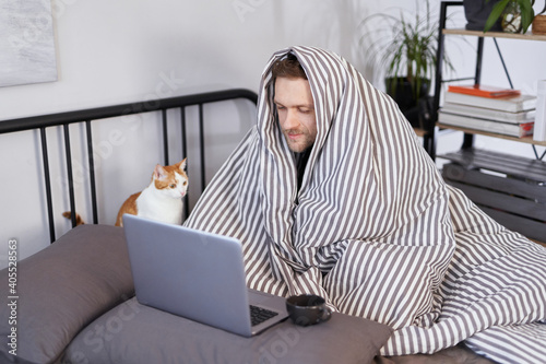Young attractive caucasian man sitting on a bed wrapped with blanked at home working using laptop with cup of hot drink. Guy browsing Internet for flu, influenza, cold and covid-19 symptoms