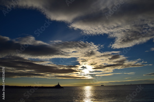 Tramonto invernale giallo sul mare con piccolo faro su diga in controluce, nuvole in cielo che coprono il sole
