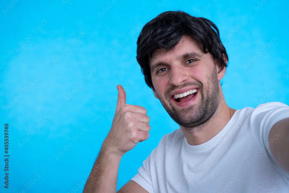 Naklejka premium Photo of handsome man in casual t-shirt and bristle on face smiling on camera with thumb up while taking selfie isolated over blue background