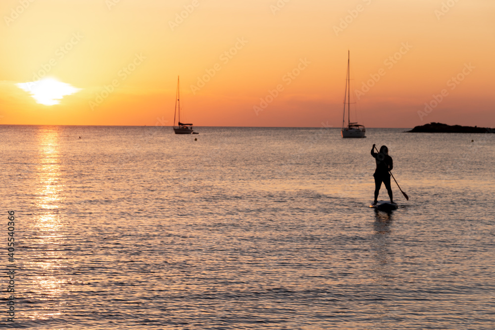 Man on stand up paddle board at sunset in calm waters with a sailboat