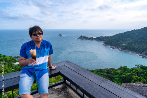 man sit at top view point watching sea coast holding latte coffee, asian man relax with coffee and ocean view  © Thitiporn