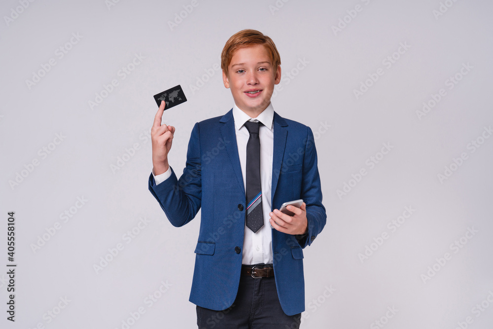 Confident young teen boy in formal outfit with red hair and braces holding smart phone and credit card isolated over grey background Confident young teen boy in formal outfit with red hair and braces holding smart phone and credit card isolated over grey background