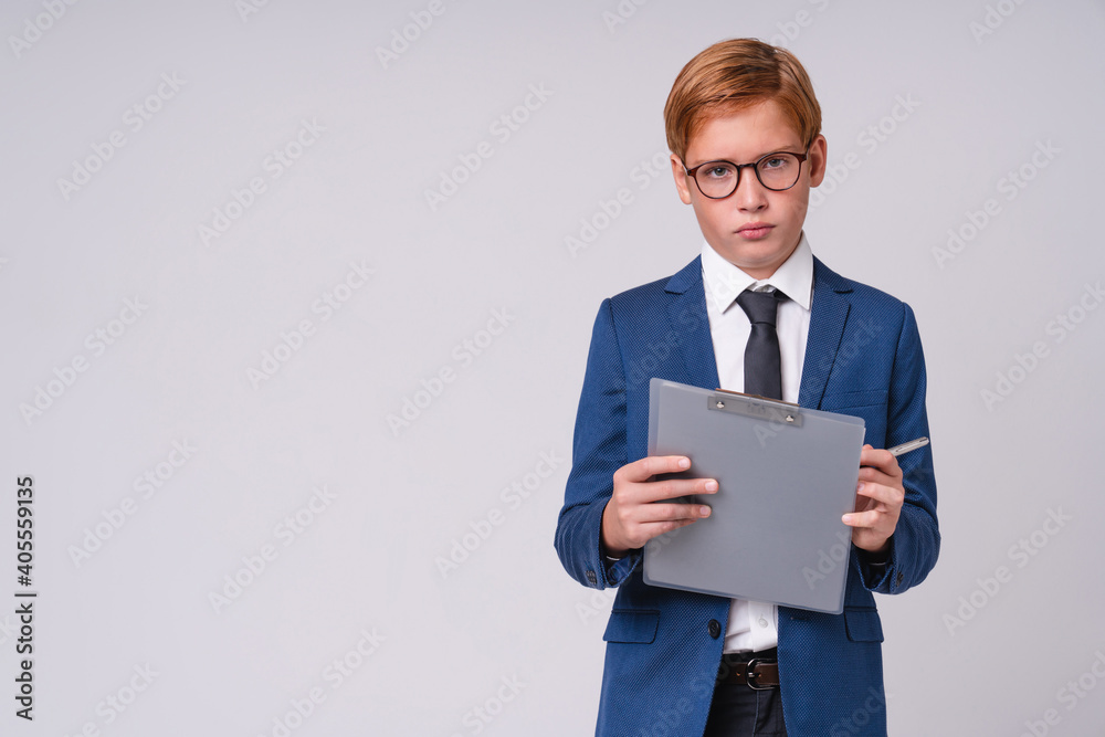 Busy young preschool redhead boy in formal suit and clipboard isolated over grey background Busy young preschool redhead boy in formal suit and clipboard isolated over grey background