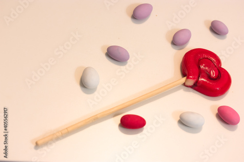 Lollipop on a stick in the shape of a red heart on a white table surrounded by colorful candy, layout. A sweet treat for Valentine's Day 